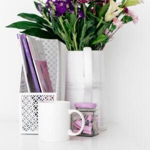 Photograph of a white mug, with a white magazine rack, pot of pink paper clips and a jug with flowers